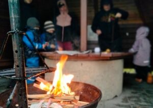 Edustalla nuotio ja takana ihmisiä nuotiokatoksessa.
